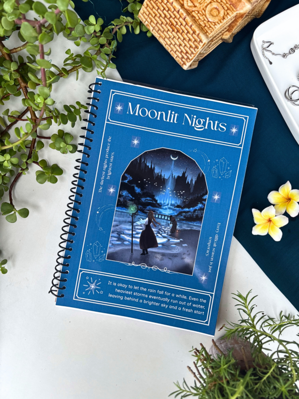 A4 spiral notebook with a midnight-blue "Moonlit Nights" cover featuring an illustrated starry forest path with a lone figure and lanterns, framed with sparkle and crystal details, and a reflective quote at the bottom.