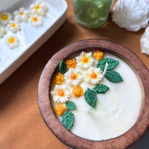 Handcrafted scented candle in a natural wooden bowl, topped with white and orange wax flowers and green leaves, with customisable wax colour and scent.