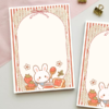 A6 notepad with a lace-bordered design featuring a cute bunny making strawberry jam toast, surrounded by strawberries and flowers, with a striped red border and bow.