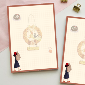 Grid notepad inspired by Kiki's Delivery Service, featuring an illustrated wreath with a black cat and a small figure with a red bow carrying a delivery bag, in warm muted tones.