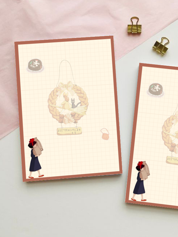 Grid notepad inspired by Kiki's Delivery Service, featuring an illustrated wreath with a black cat and a small figure with a red bow carrying a delivery bag, in warm muted tones.